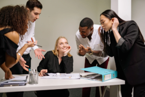 Collègues en discussion animée autour d'un bureau, une femme assise écoute en souriant